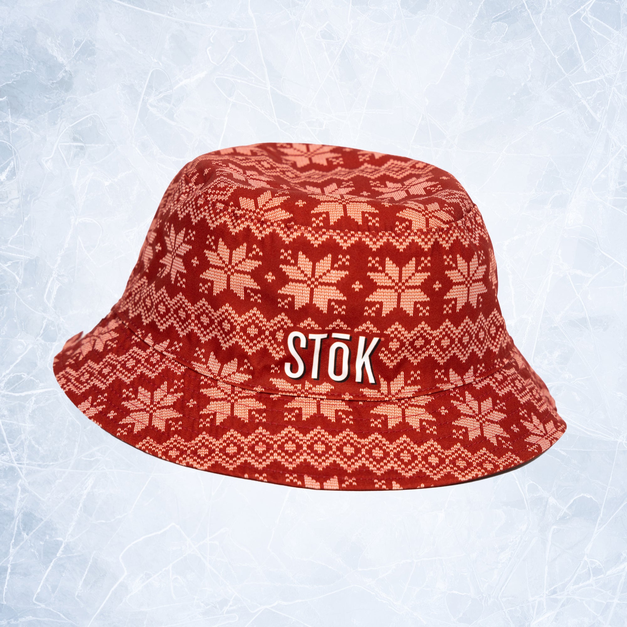 The alternate reversible inside of the red bucket hat flipped out with a festive, knitted pattern design featuring white snowflakes and geometric shapes throughout. The Stōk logo is displayed in white on the front of the hat, maintaining a clean and simple contrast against the intricate design.