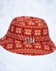 The alternate reversible inside of the red bucket hat flipped out with a festive, knitted pattern design featuring white snowflakes and geometric shapes throughout. The Stōk logo is displayed in white on the front of the hat, maintaining a clean and simple contrast against the intricate design.