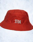 A red bucket hat featuring the Stōk logo in white lettering on the front. The image hints at the other side of the bucket hat resembling a festive, winter theme.