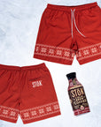 The front and back of red swim trunks with a strip of knitted pattern design featuring white snowflakes and geometric shapes across the bottom of the legs. The Stōk logo is displayed in white on the left leg strip of pattern, maintaining a clean and simple contrast against the intricate design. There is a white shoelace like tie closure on the front of the waistband next to a Stōk cold brew