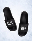 Black rubber slides with the white Stōk logo written across the strap of both.