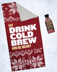 A red towel for Stōk cold brew coffee featuring a holiday-themed design. The sign has a red background with white knitted patterns resembling snowflakes and festive decorations. The large text in the center reads ‘EAT DRINK COLD BREW AND BE MERRY,’ with ‘Stōk’ logo displayed at the bottom next to a Stōk Cold Brew