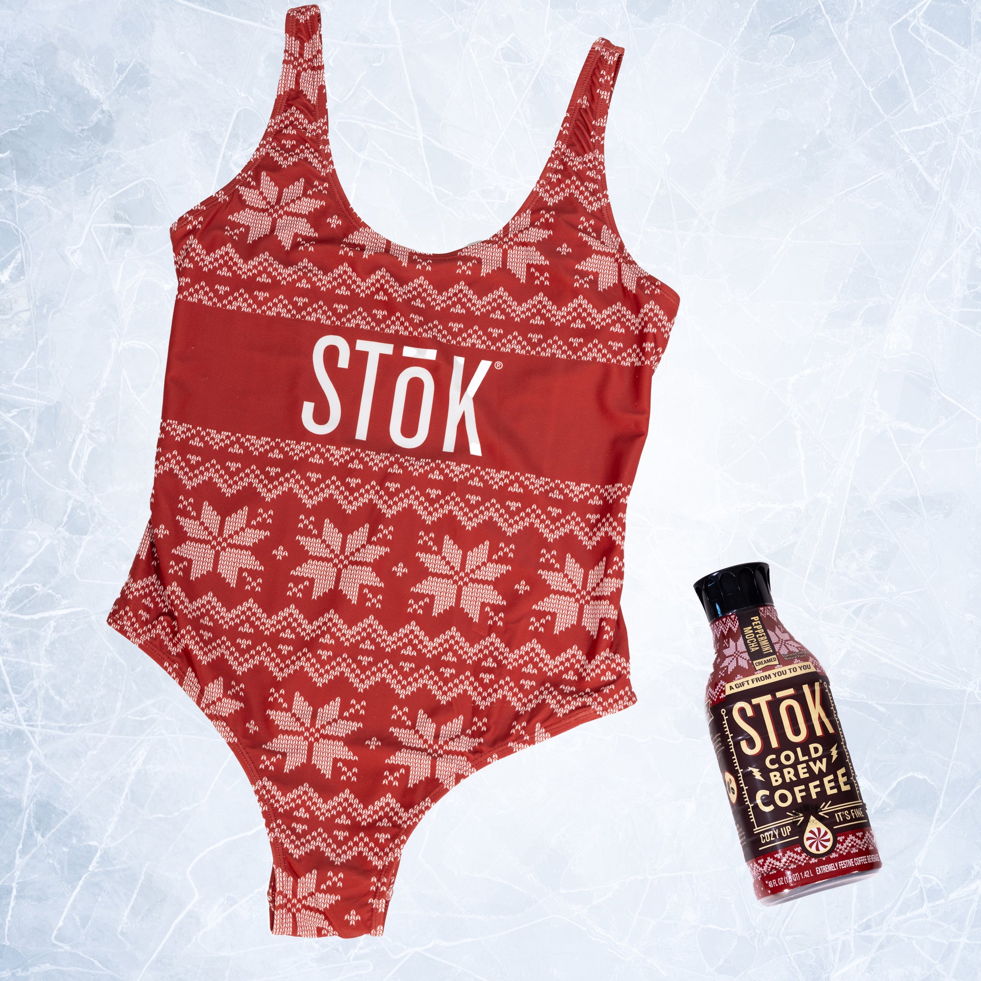 The front of a red one piece swimsuit with a knitted pattern design featuring white snowflakes and geometric shapes throughout. The Stōk logo is displayed in white on the front across the chest, maintaining a clean and simple contrast against the intricate design next to a Stōk cold brew.