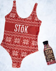 The front of a red one piece swimsuit with a knitted pattern design featuring white snowflakes and geometric shapes throughout. The Stōk logo is displayed in white on the front across the chest, maintaining a clean and simple contrast against the intricate design next to a Stōk cold brew.